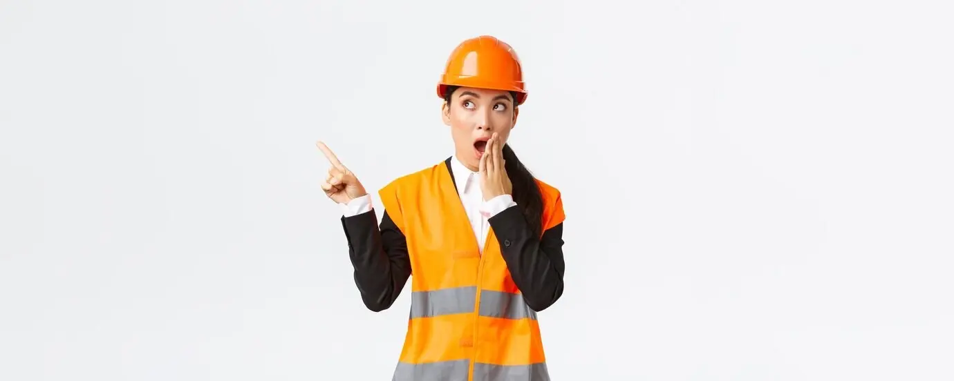 Mujer asiática, ingeniera y gerente de construcción, con casco de seguridad y chaleco reflectante, boquiabierta, señalando con el dedo hacia la esquina superior izquierda, conmocionada e impresionada, asombrada al descubrir algo interesante.